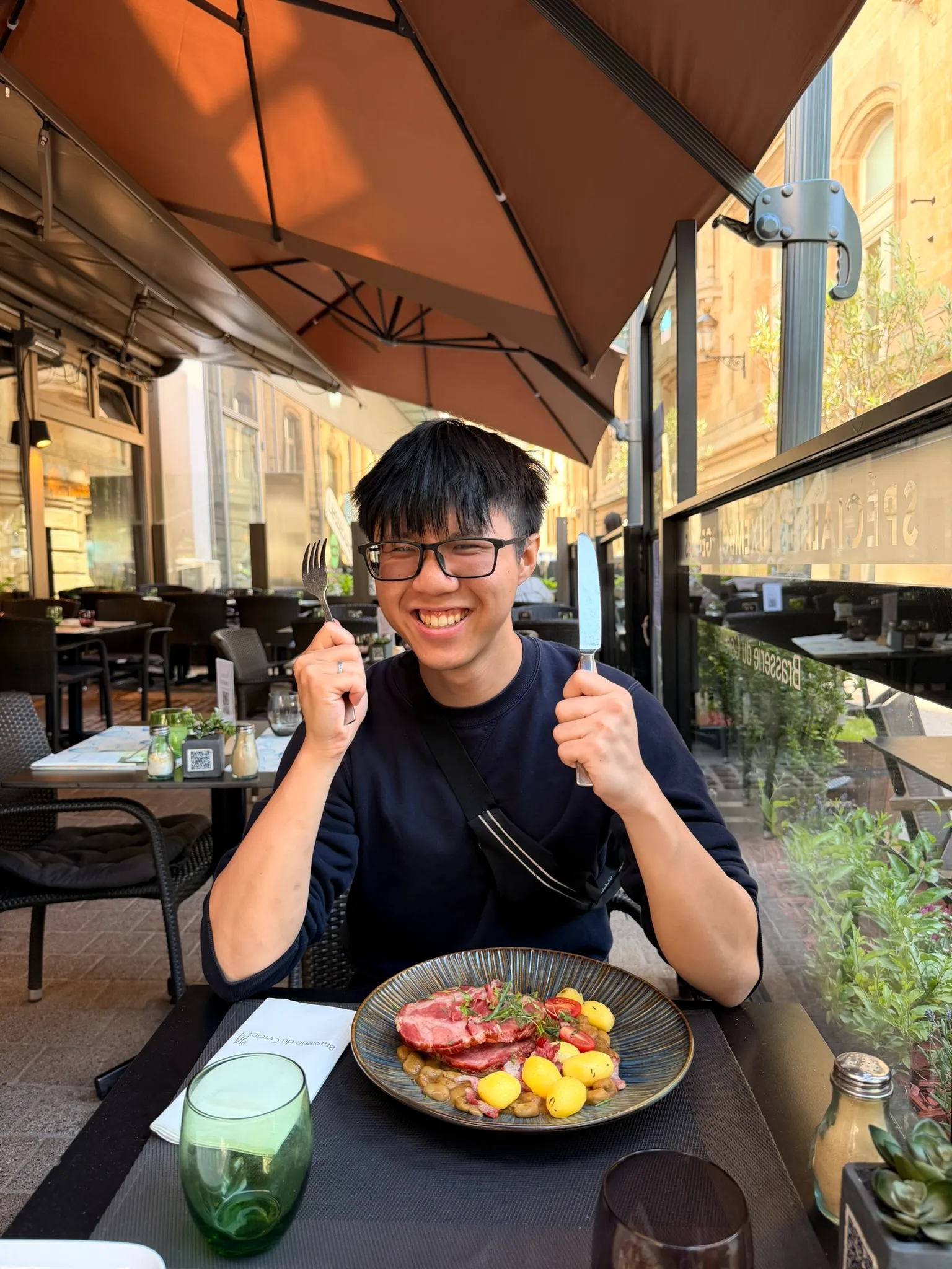 Sean grinning with a fork and knife raised over a plate of pork, beans and potatoes on a brasserie terrace